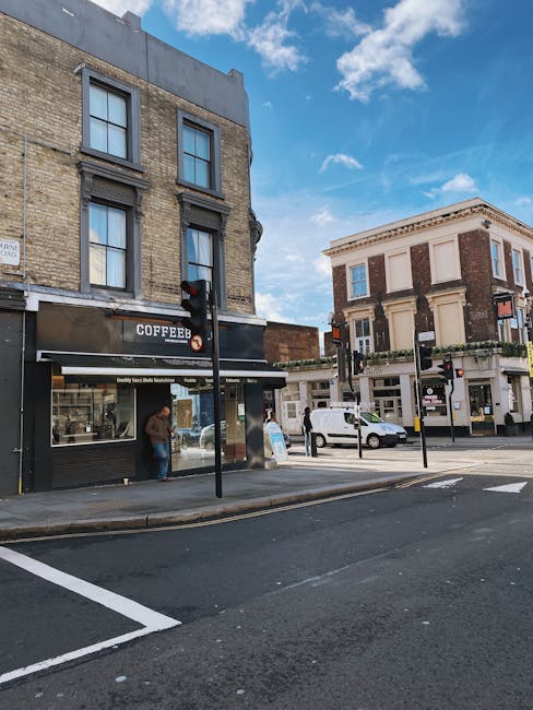 A street scene outside a building on Anerley Road, showing a black and grey coffee shop storefront with large windows, a person standing near the entrance, and another person walking past. In front of the shop, there is a traffic light and a small paved area with pedestrian crossings, bollards, and parking spaces. Nearby, a white van is parked on the street, adjacent to a multi-storey three-storey brick building with white window frames and decorative architectural details. The sky above is partly cloudy with patches of blue, and the environment suggests an urban area suitable for house or office removals, including furniture transport and loading processes involving nearby vehicles and pedestrian pathways.