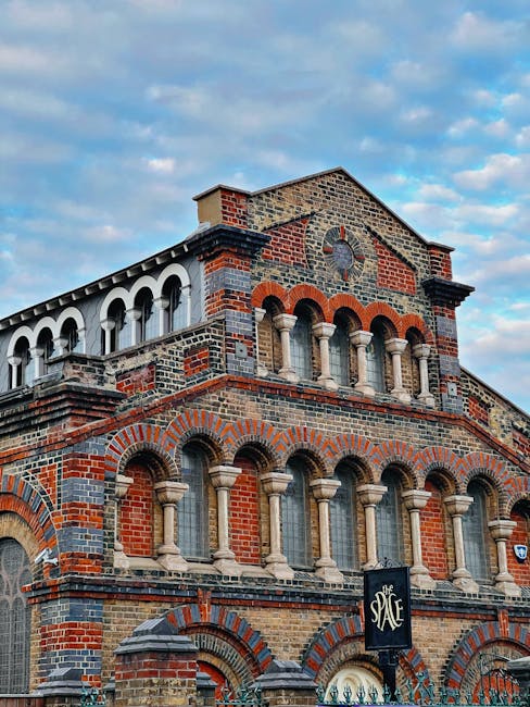 The image shows a multi-storey brick building with a combination of red and grey brickwork, featuring arched windows with stone mullions and decorative brick patterns. Small columns support the upper windows, and a large clock face is visible near the top part of the facade, beneath a gabled roof. The sky above is partly cloudy with patches of blue, and a black sign with white text reading 'The SPACE' hangs from a metal bracket attached to the building. This structure appears to be an older, historic property, potentially used as a commercial or community space in an urban setting. Inside, there are no visible furniture or moving equipment, indicating no active moving process, but the building's detailed architecture emphasizes its historic character. The scene is well-lit with natural daylight, highlighting the intricate brickwork and architectural features, making it relevant for topics related to building renovations, historic property management, or urban development.