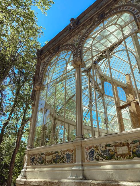 Photograph of a grand, vintage glass conservatory with intricate metal framing and decorative architectural details, located outdoors with visible trees in the background. The conservatory features la