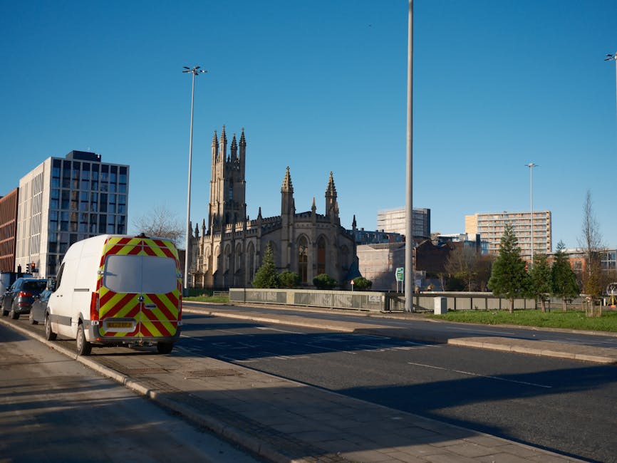 Street view showing a white commercial van with red and yellow chevron markings parked on the side of a road near a sidewalk; behind the van, there is a historic Gothic-style church with tall spires and intricate detailing, surrounded by modern office buildings and trees. The scene is illuminated by bright sunlight with a clear blue sky overhead, and streetlights are visible along the pavement. The image depicts an urban area in Crystal Palace, with the church as a notable landmark, and the van is positioned to facilitate home relocation or furniture transport services in line with house removals. The setting suggests a busy city environment suitable for loading or unloading furniture and boxes during a house move, as handled by companies like [COMPANY_NAME] specialized in removals.
