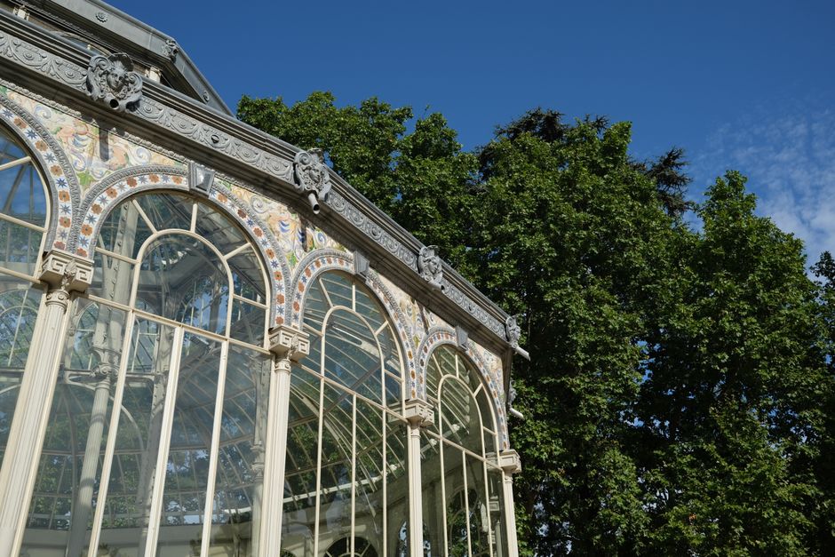 A detailed exterior view of a historic glass conservatory or greenhouse featuring ornate white metal framing with decorative scrollwork and floral motifs. The structure has large, arched windows fitted with multiple panes of glass, supported by vertical and horizontal bars, allowing natural light to illuminate the interior. The building is situated outdoors, with lush green trees and a bright blue sky with scattered clouds visible in the background, indicating a clear day. This scene is relevant to home relocation or furniture transport, as it depicts a delicate glass structure that would require professional removals services for safe packing and transportation, as offered by companies like removal companies in Crystal Palace, especially during house moves from Upper Norwood to new locations.