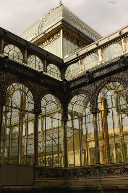 Exterior view of an ornate, historic glasshouse or conservatory with decorative metal framework, large arched windows, and intricate ironwork details. The structure features multiple glass panels, som