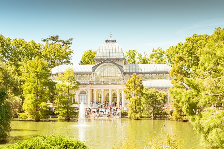 Exterior view of an ornate, historic glasshouse or conservatory with decorative metal framework, large arched windows, and intricate ironwork details. The structure features multiple glass panels, som