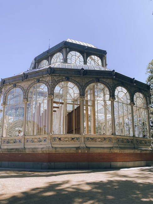 A large, ornate greenhouse structure with a decorative metal frame and numerous glass panels situated outdoors on a paved surface, partially shaded by nearby trees. The greenhouse features a domed roof with additional glass sections and decorative detailing along the lower edges. Inside the greenhouse, visible through the glass panels, are metal shelving units and potted plants, suggesting ongoing maintenance or cleaning. The setting appears to be during daylight with clear skies, creating natural light that reflects off the glass surfaces. This image exemplifies the type of property feature or structural element that might be involved in a house removal or relocation process, as handled by professional removals specialists like [COMPANY_NAME], highlighting the need for careful packing and transportation during home or furniture moves, especially when dealing with delicate glass and decorative items.